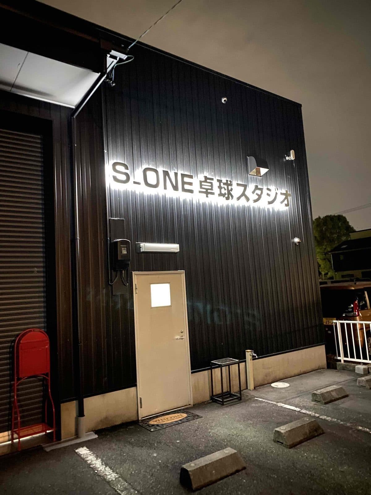 夜に看板が光るS_ONE卓球スタジオの入口。黒い外壁にクリーム色の扉、手前に駐車場